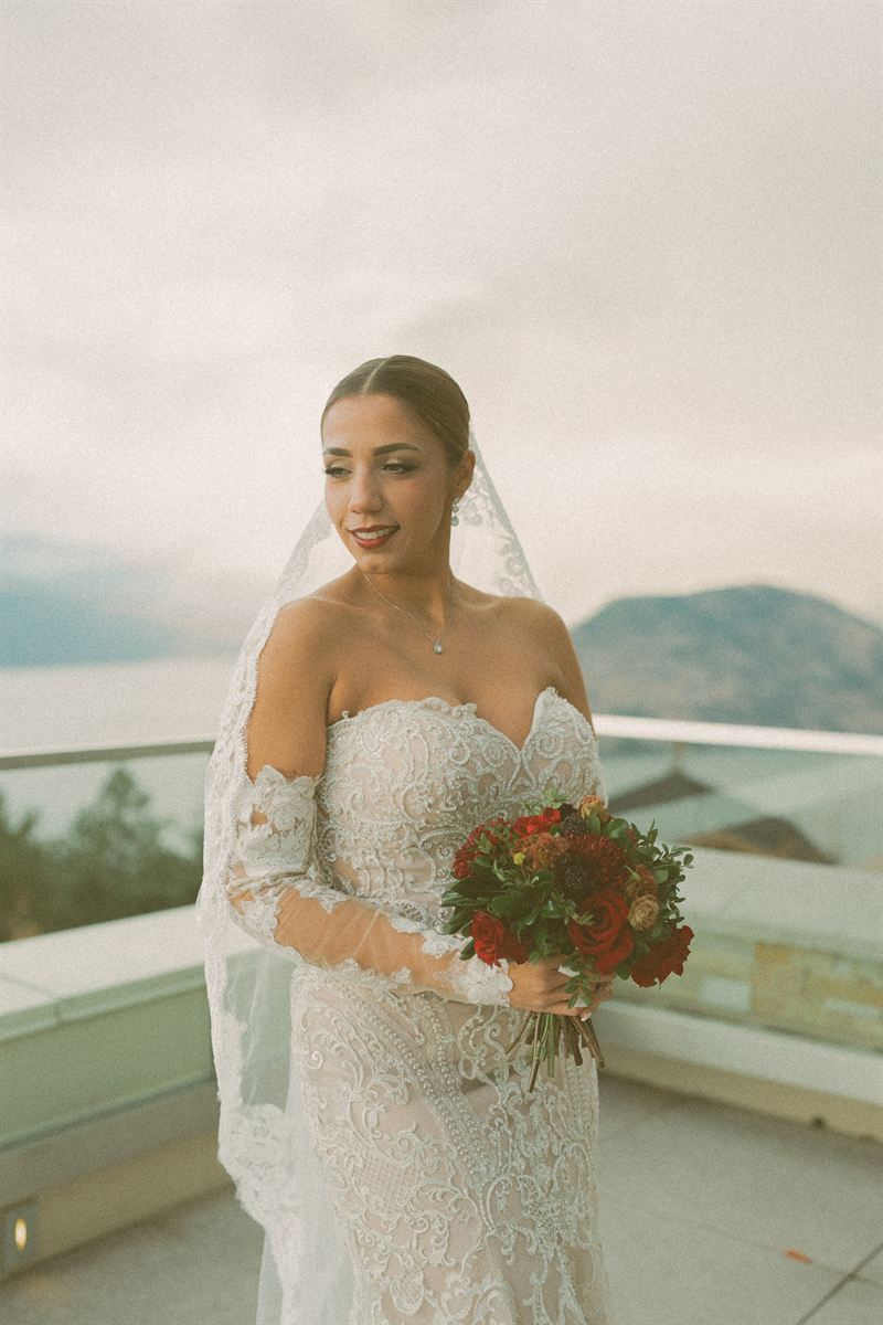 Bride on a terrace above Okanagan Lake in a lace gown and cathedral veil, holding a deep red autumn bouquet