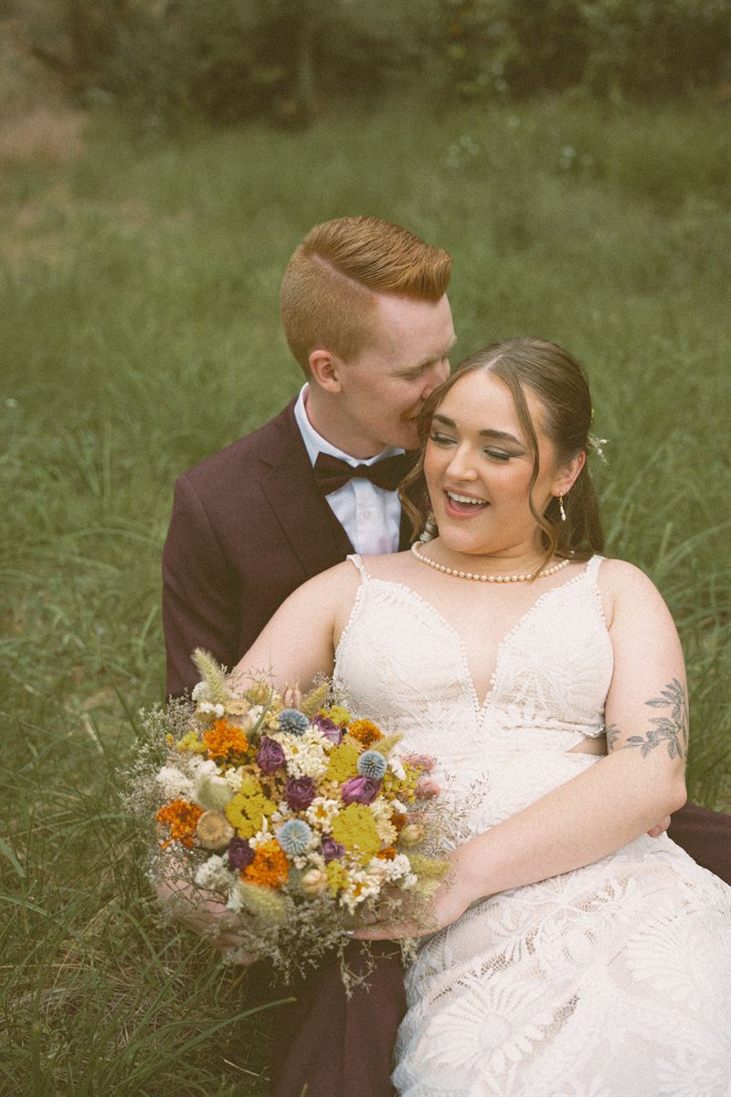 Groom kissing the bride's cheek as she laughs, the two of them sitting in a summer meadow